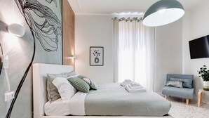 Ein modernes Schlafzimmer mit einem Bett, einem Nachttisch, einem Sessel, einem an der Wand montierten Fernseher und einem Wandbild.