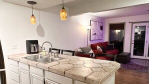 Una cocina con una isla central, dos lámparas colgantes, un fregadero y una zona de comedor con un sofá y sillas rojas.