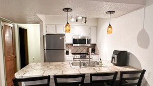 Cocina compacta con una isla central, armarios blancos, electrodomésticos de acero inoxidable e iluminación colgante.