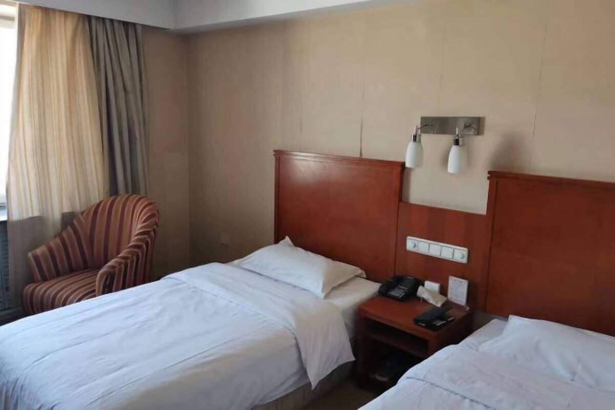 Hotellihuone, jossa on kaksi sänkyä, raidallinen nojatuoli, yöpöytä, jossa on puhelin, ja seinälle asennettu valaisin.