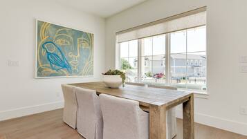 A modern dining area with a wooden table and cushioned chairs, a large abstract painting on the wall, and a large window with a view of the neighborhood.