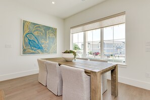 A modern dining area with a wooden table and cushioned chairs, a large abstract painting on the wall, and a large window with a view of the neighborhood.