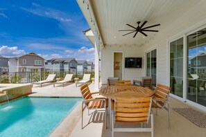 A poolside area with a wooden table and chairs, a ceiling fan, and a view of neighboring houses.