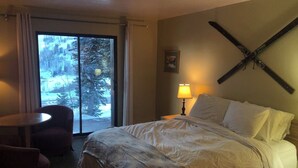 Habitació d'hotel amb llit, una cadira, una taula i una finestra amb vista a arbres coberts de neu.