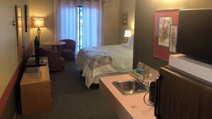 Habitació d'hotel amb un llit, una escriptori amb una làmpada, una cadira, una taula petita, un lavabo i una televisió.