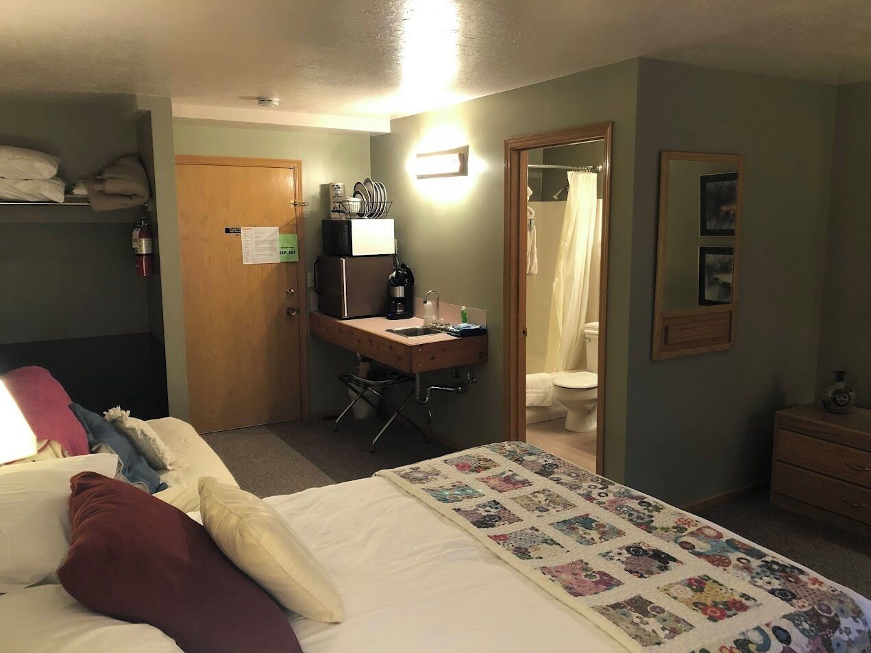 A hotel room with a bed, a desk with a computer, a chair, a bathroom with a shower curtain, and a small dresser.