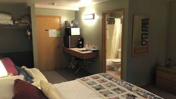 A hotel room with a bed, a desk with a computer, a chair, a bathroom with a shower curtain, and a small dresser.