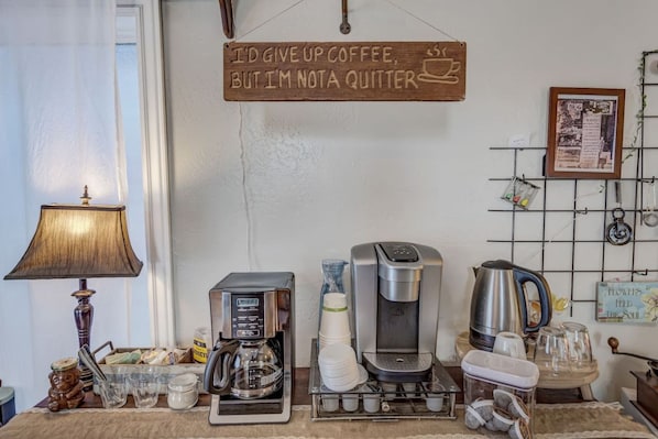 A coffee station with a coffee maker, cups, and a sign saying 'I'D GIVE UP COFFEE, BUT I'M NOT A QUITTER.'
