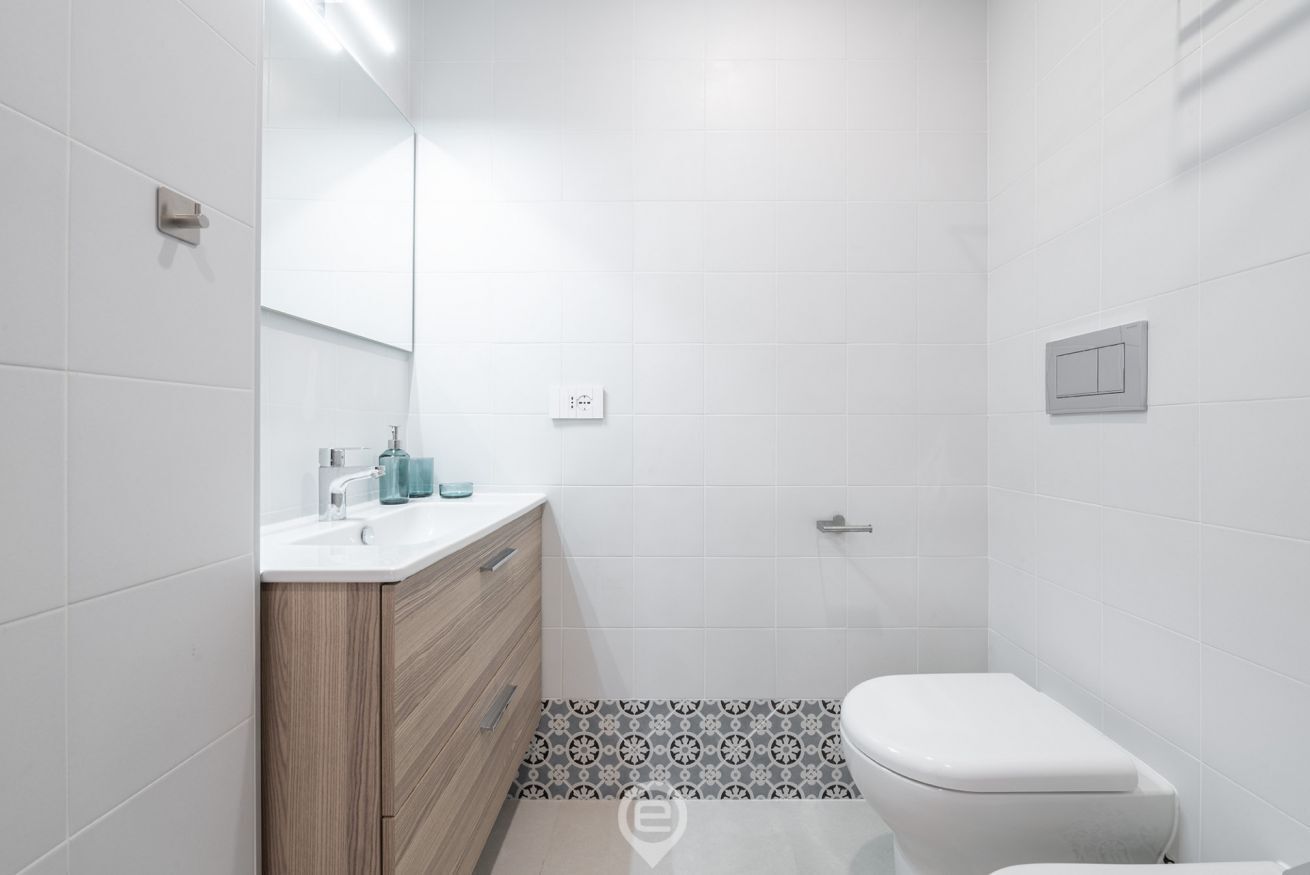Un baño moderno con inodoro blanco, tocador de madera con espejo y suelo estampado.