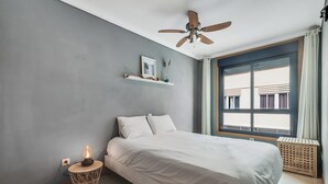 A bedroom with a bed, a ceiling fan, a window with curtains, a bedside table with a lamp, and a wall-mounted shelf with decorative items. - Home2Book Trendy Apt in Las Palmas Center (Las Palmas de Gran Canaria)