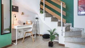 A modern room with a white desk, a chair, a lamp, a mirror, a small shelf with decorative items, a potted plant, a black bag, and a staircase with wooden steps. - Home2Book Trendy Apt in Las Palmas Center (Las Palmas de Gran Canaria)