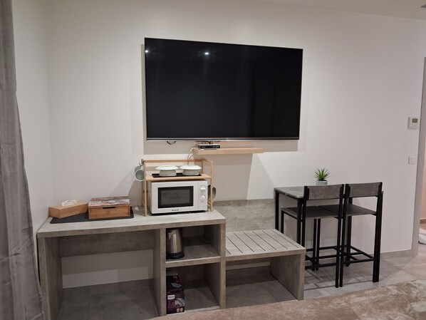 Un espacio habitable compacto con un televisor de pantalla plana montado en la pared, una pequeña mesa de madera con un microondas, una tetera y una caja de pañuelos. Hay una sola silla negra y una pequeña planta en maceta.