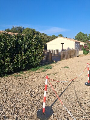 Ein von einer Hecke umgebener Bereich mit einem Kiesweg, der zu einem Gebäude mit sandfarbener Fassade und einer Satellitenschüssel führt.