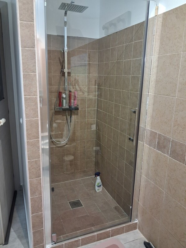 Box doccia con pareti in vetro, soffione a mano, piccolo ripiano con articoli da toilette e scarico a pavimento.