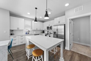 Une cuisine moderne avec des meubles blancs, un îlot central doté de tabourets, un four à micro-ondes, un réfrigérateur et des luminaires suspendus.
