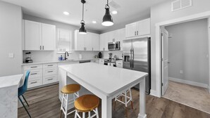 Cozinha moderna com armários brancos, uma ilha central com banquetas, micro-ondas, geladeira e iluminação pendente.