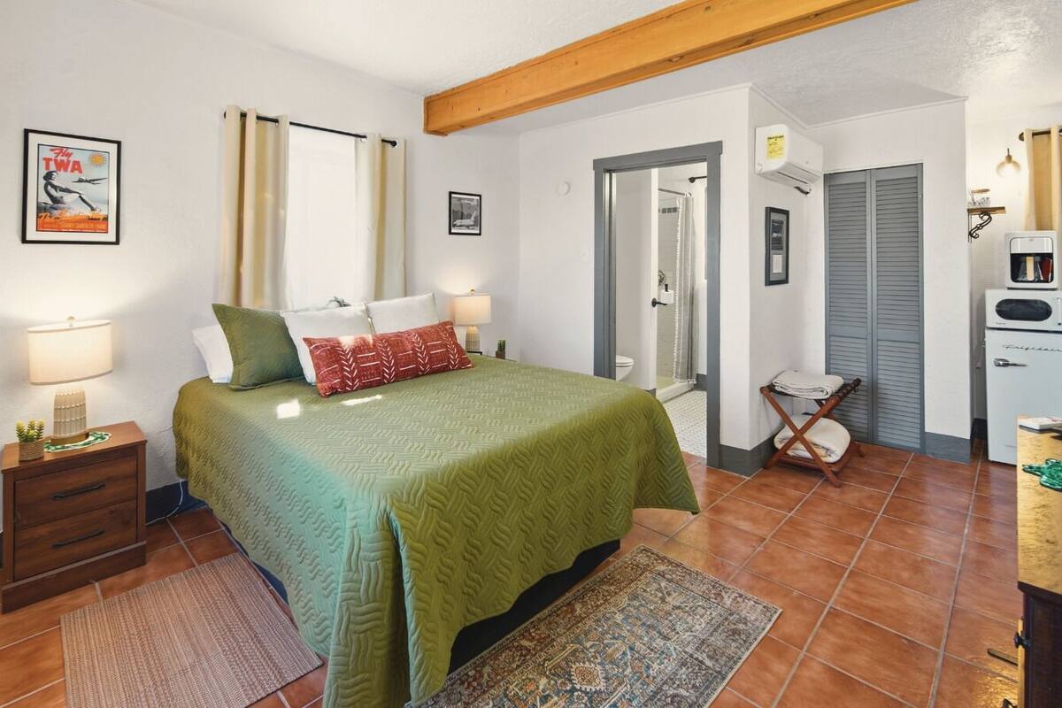 A bedroom with a bed, bedside tables, a nightstand, a mirror, a wardrobe, a radiator, and a small kitchenette.