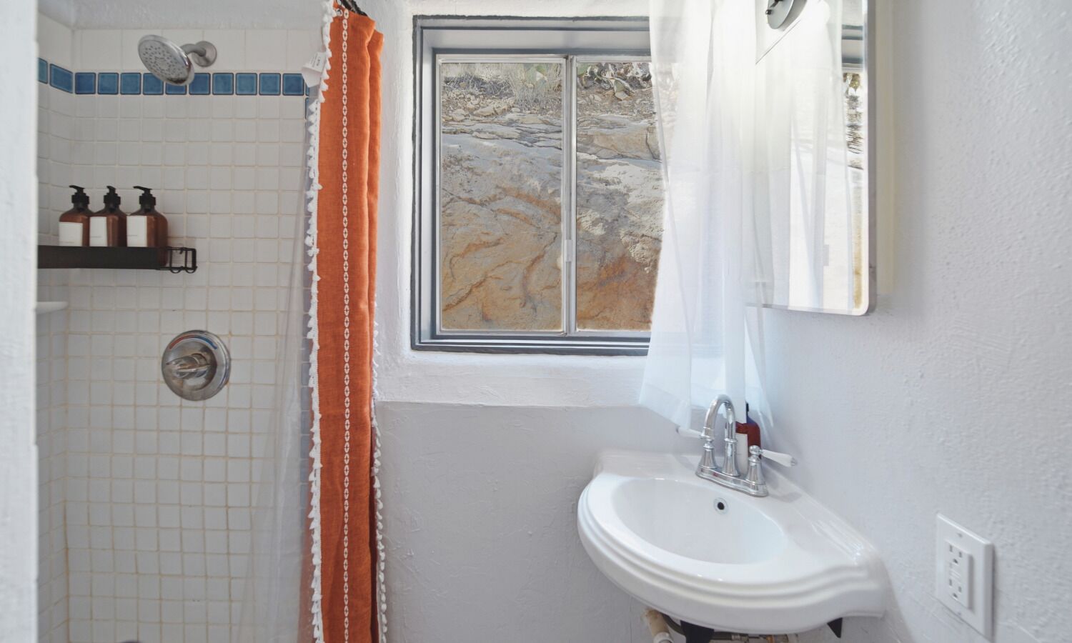 A bathroom with a shower, a sink, a window with a view of a rocky landscape, and a towel rack with bottles.