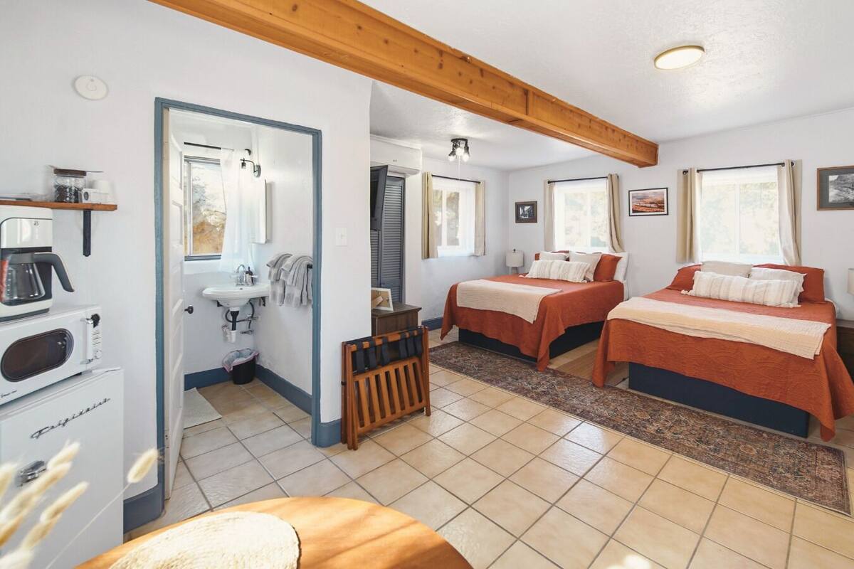 A compact hotel room with two beds, a kitchenette, a bathroom, and a small sitting area.