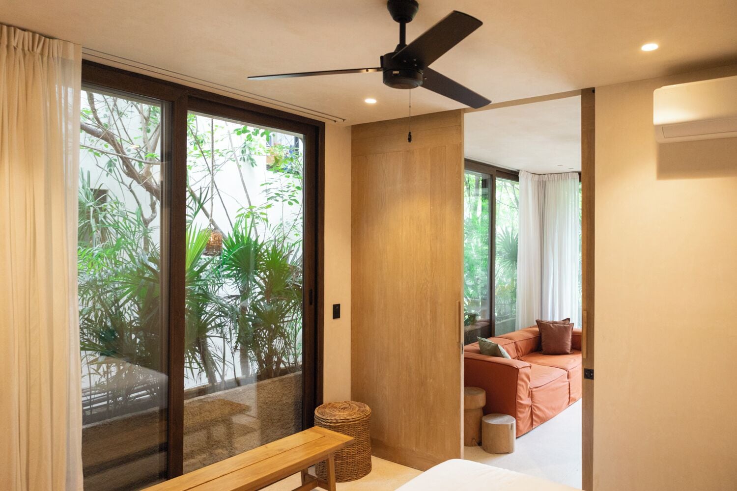 Une chambre moderne dotée d’une porte-fenêtre coulissante donnant sur un balcon, d’un ventilateur de plafond, d’un banc et d’un canapé avec des coussins.
