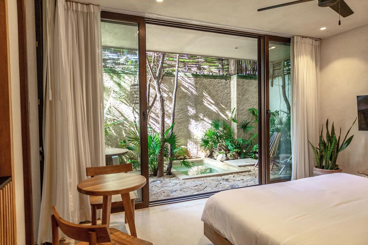 A bedroom with a bed, a wooden chair, a small table, and a sliding glass door leading to a pool area with plants.