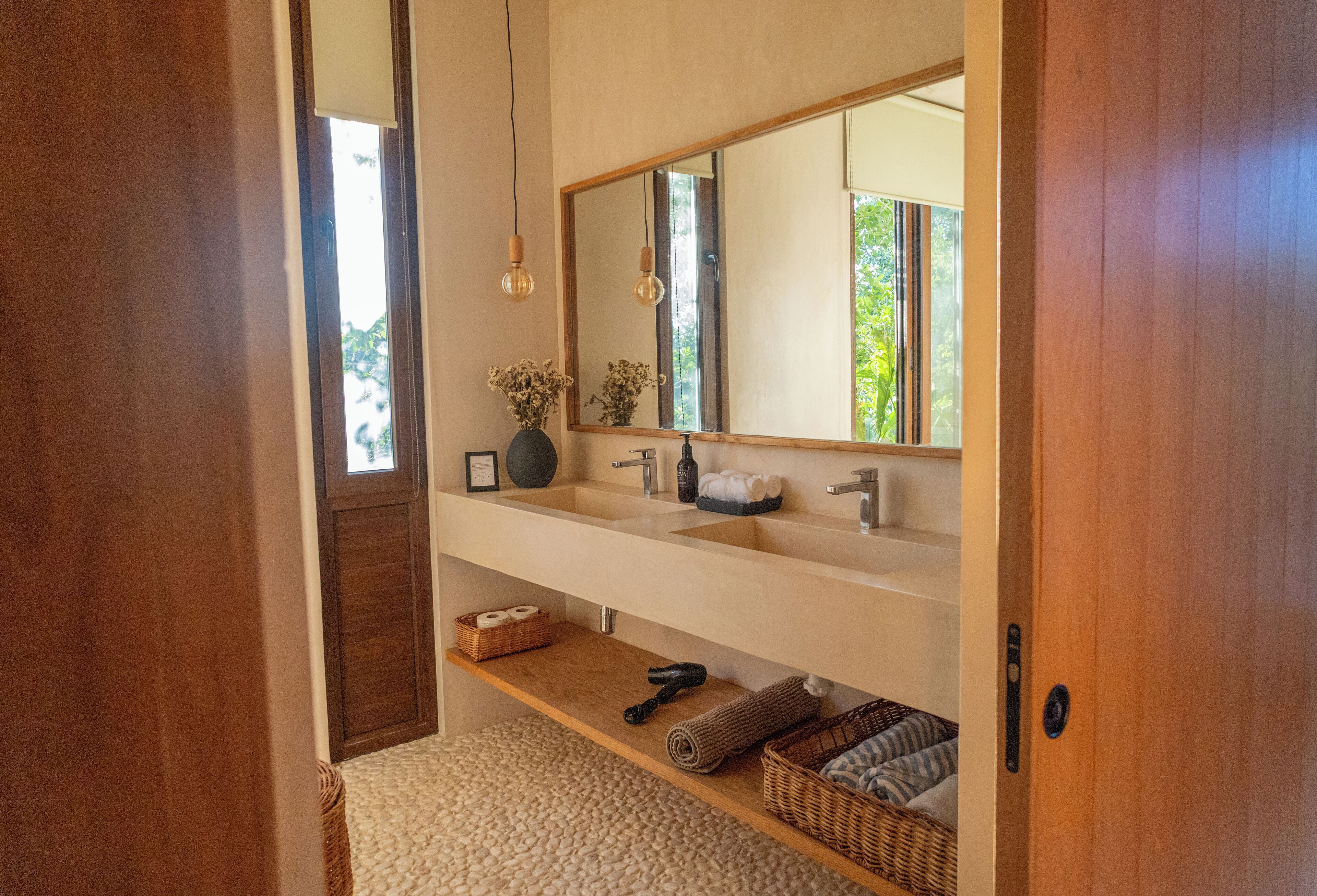 A bathroom with a wooden door, a large mirror, two sinks, a towel rack, a shelf with toiletries, and a window with natural light.