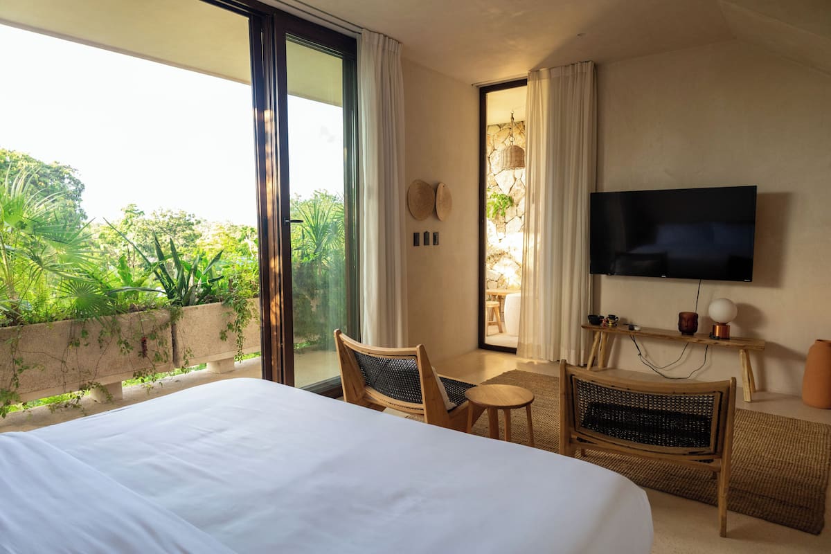 A hotel room with a large bed, a television, a wicker chair, a small table, and a view of greenery outside.