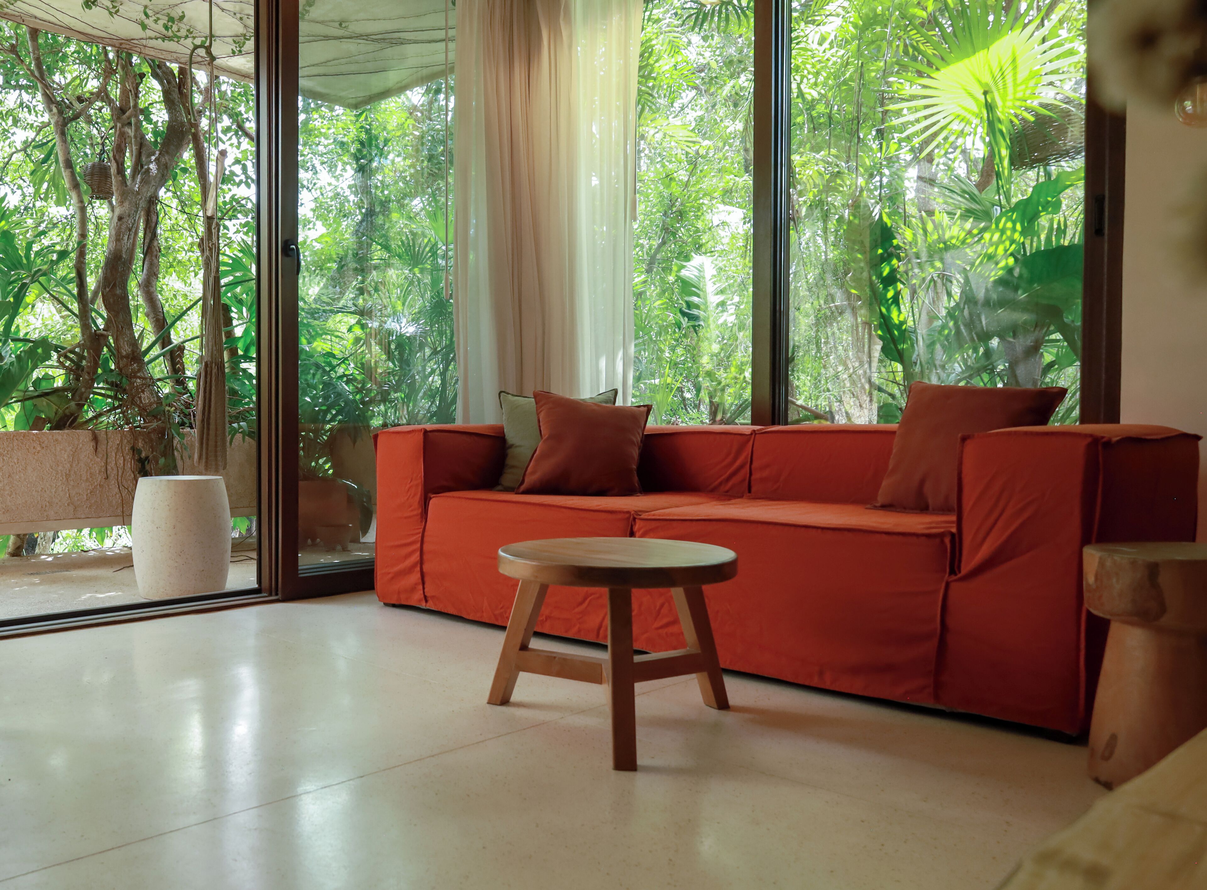 A modern living room with a red sofa, a round wooden table, and large windows offering a view of lush greenery.