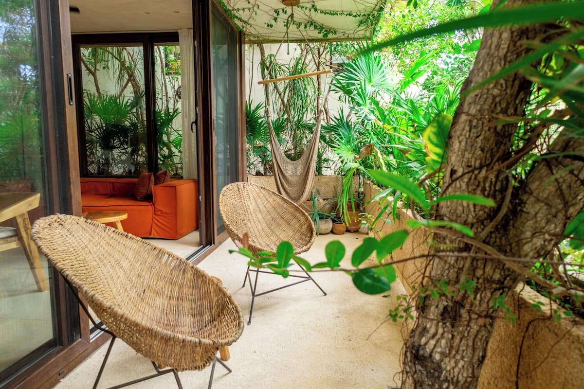 A room with wicker chairs, a hanging chair, and a sofa with orange cushions.