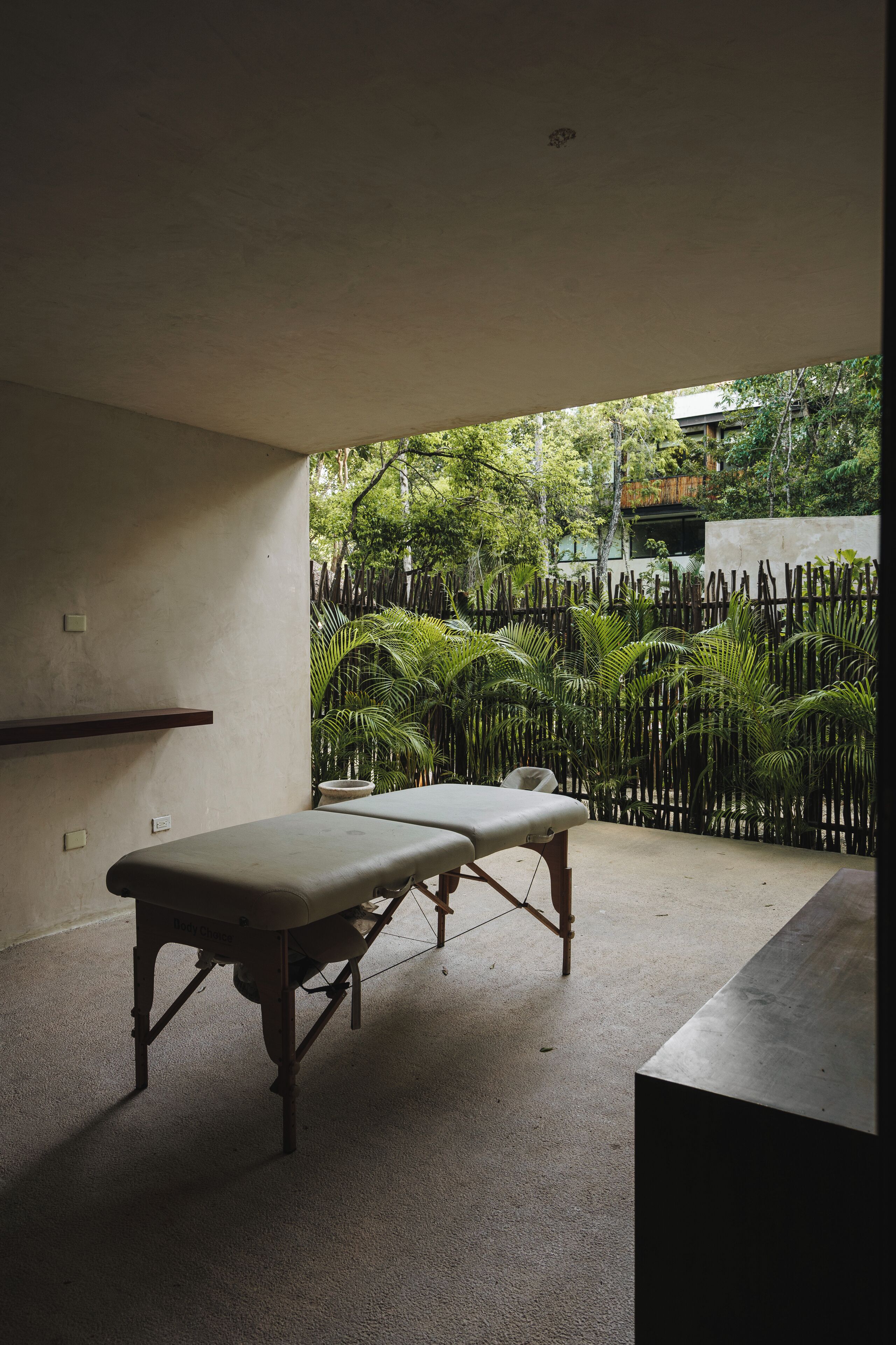 Une salle de soins équipée d’une table en bois et d’un banc, avec un sol en béton et une vue sur une clôture en bambou et la verdure environnante.