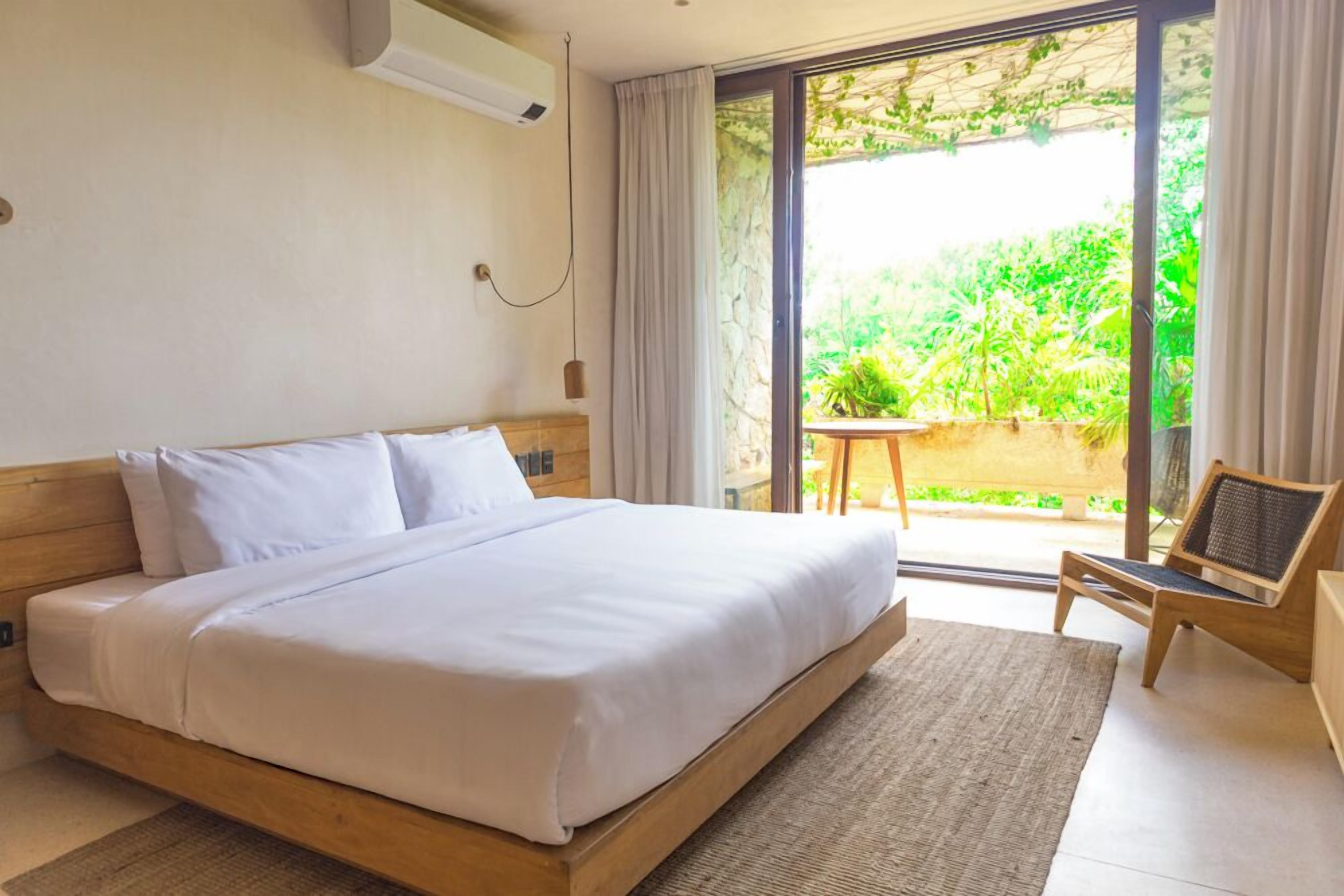 Une chambre avec un grand lit, une tête de lit en bois, une petite table, une chaise et une vue sur la verdure grâce à une porte-fenêtre coulissante.