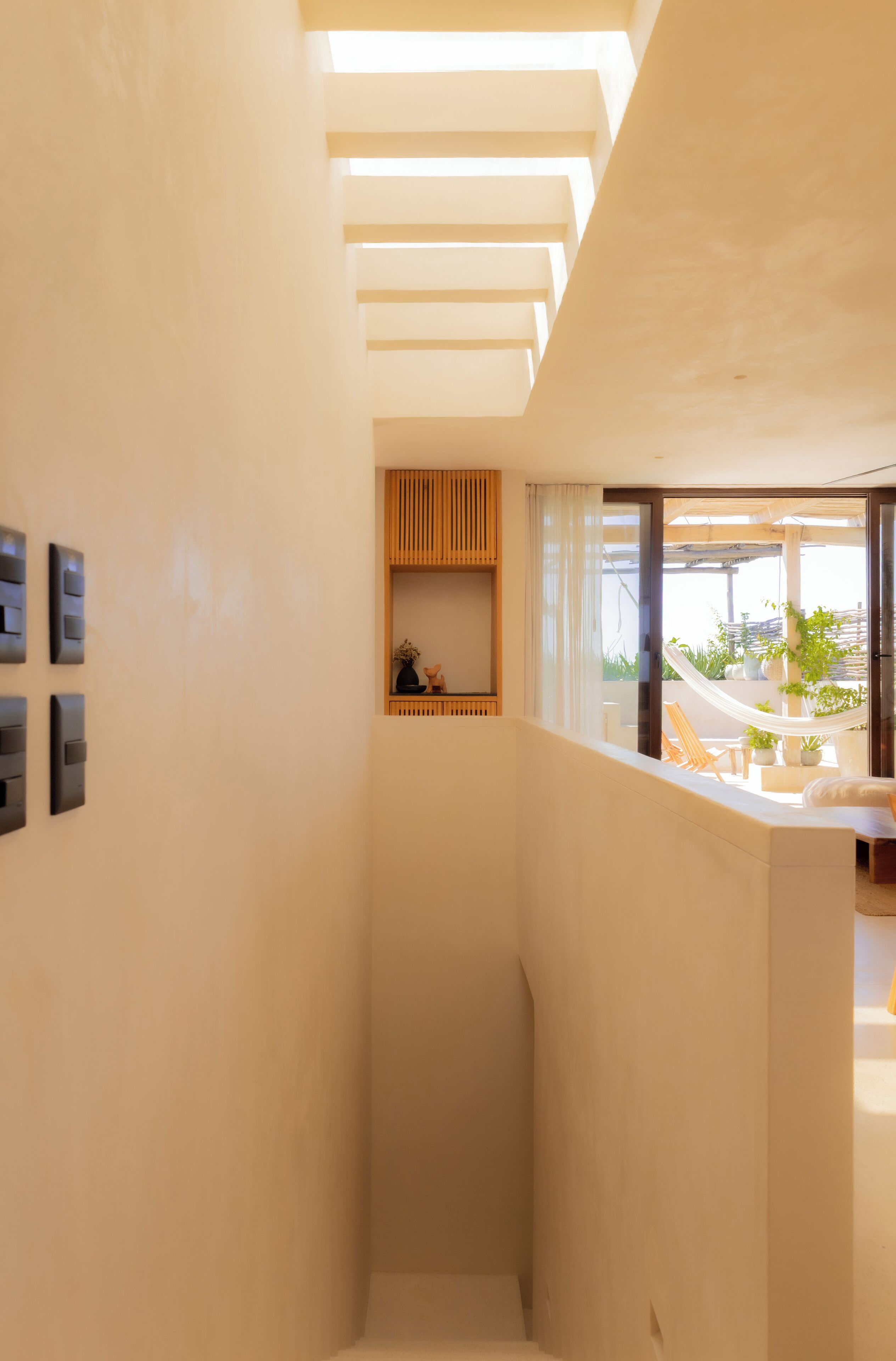 Un intérieur moderne avec une colonne centrale, une étagère en bois agrémentée d’objets décoratifs et une vue sur un espace extérieur avec des plantes et un hamac.