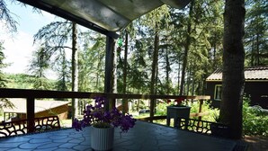 A covered outdoor area with a stone floor and a wooden railing. There is a potted plant with purple flowers on a table. - Chalet avec piscine au Domaine du Lac de Goulens (LAYRAC)