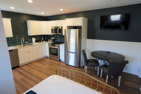A compact kitchen with white cabinets, stainless steel appliances, a microwave, oven, and refrigerator. There's a dining table with chairs and a TV mounted on the wall.