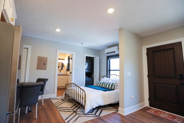 A bedroom with a bed, a desk, and a chair. There is a door and an air conditioning unit on the wall.