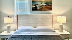 Una camera da letto con un letto, due comodini con lampade, una finestra con persiane e un quadro raffigurante una spiaggia.