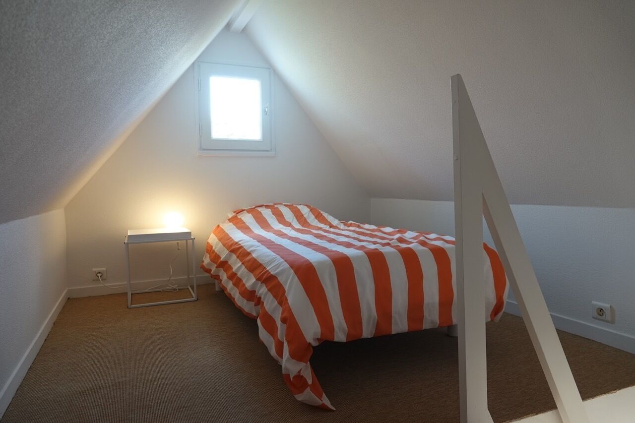A small, triangular attic bedroom with a sloped ceiling, a single bed with an orange and white striped bedspread, a small bedside table with a lamp, and a window providing natural light.