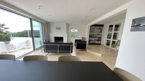 A modern living room with a black sofa, a dining table, and a fireplace. There is a bookshelf and a large window with a view of greenery. - Villa "La Loubine" contemporary thatched cottage facing the sea (Batz-sur-Mer)