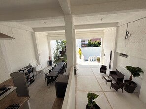 Un grand espace intérieur comprenant une longue table et des chaises, un coin cuisine et une vue sur un jardin agrémenté de plantes et d’un escalier.
