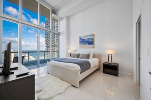 Ein modernes Schlafzimmer mit einem großen Bett, einem Schreibtisch mit Computer und Blick auf die Stadt durch große Fenster.