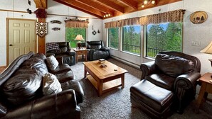 A cozy living room with leather furniture, a wooden coffee table, and large windows offering a view of greenery. - Rustic Cabin with Modern Comforts! (Cascade)