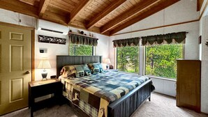 A bedroom with a large bed, wooden ceiling beams, a window with a view of greenery, a bedside table with a lamp, and a wardrobe. - Rustic Cabin with Modern Comforts! (Cascade)