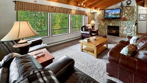 A cozy living room with leather sofas, a fireplace, a television, and large windows offering a view of greenery. - Rustic Cabin with Modern Comforts! (Cascade)