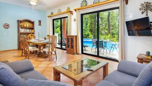 Un salon comprenant un canapé, une table basse, un coin repas avec des chaises et une table, et une vue sur une piscine, visible à travers de grandes portes-fenêtres coulissantes.