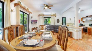 Un coin repas avec une table en bois dressée pour quatre personnes, entourée de chaises en bois. Un ventilateur de plafond est visible au-dessus, et on aperçoit une cuisine au fond.