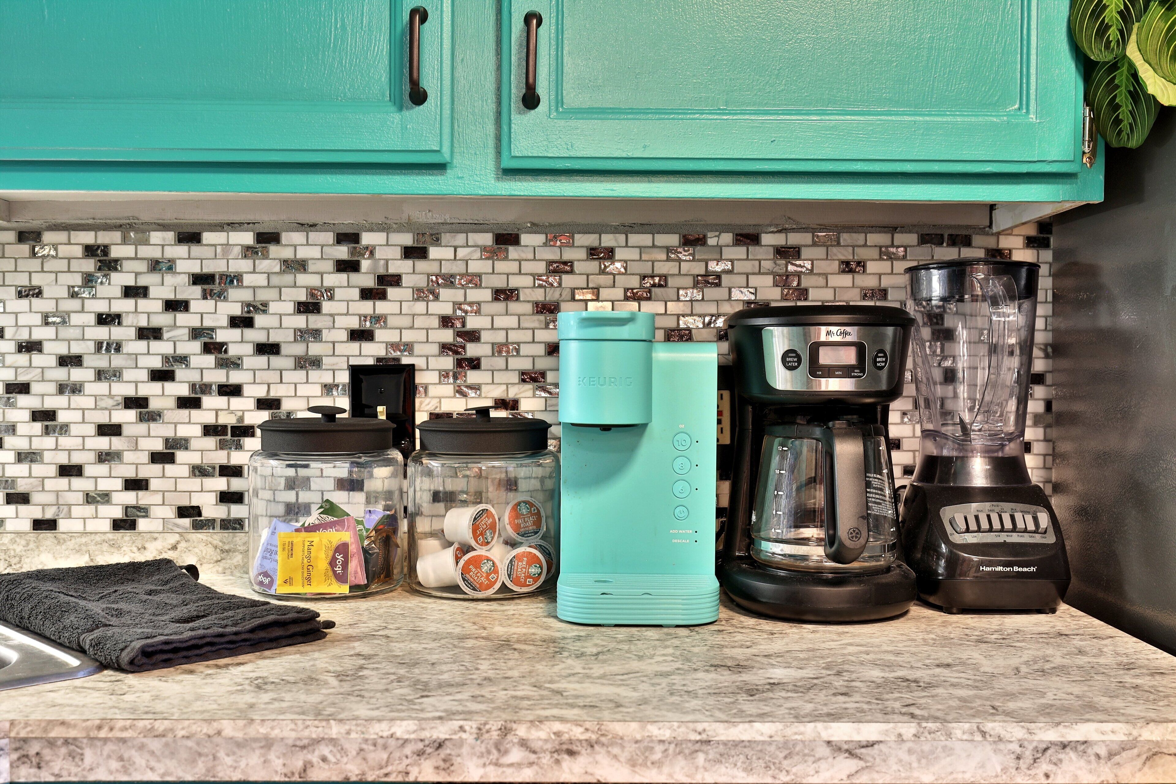 A kitchen counter with a coffee maker, a blender, and a jar of coffee pods.