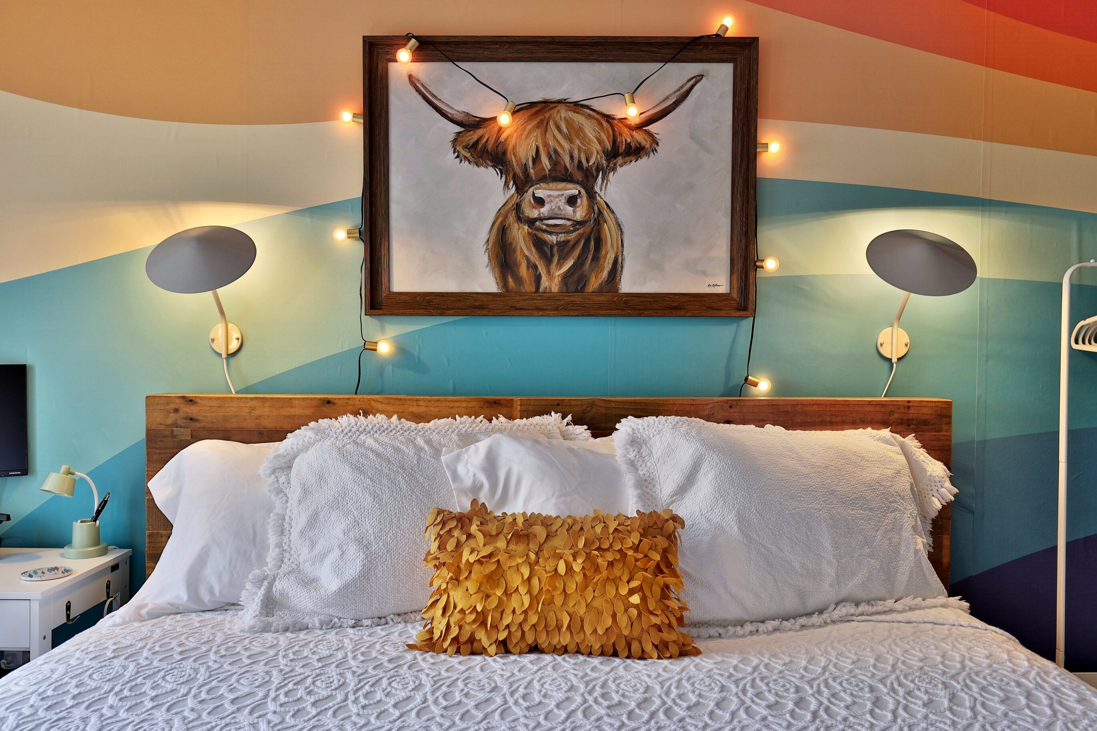 A bedroom with a wooden headboard, white bedding, a unique leaf-patterned pillow, and a framed artwork of a bull's head.