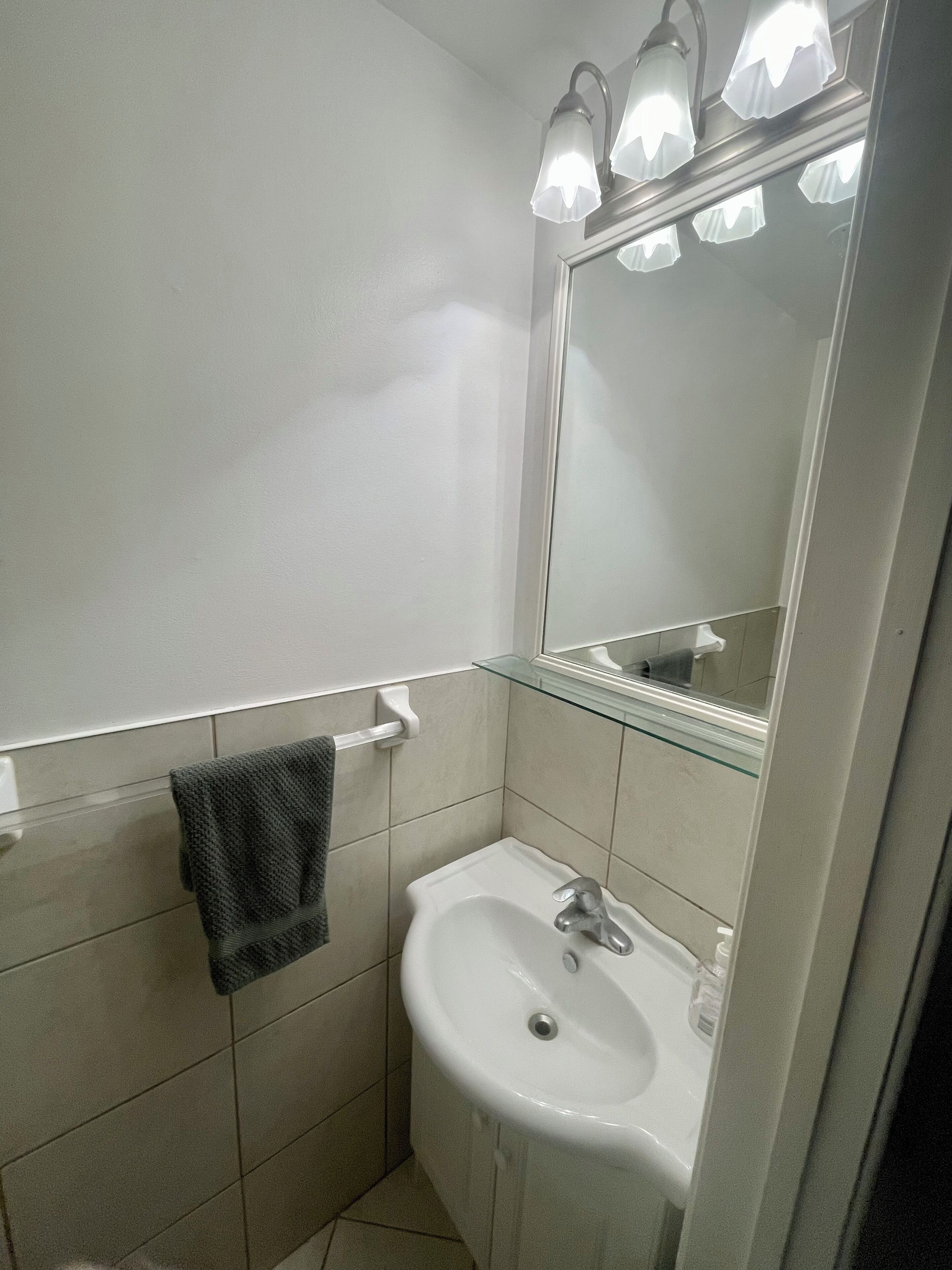 Un baño con un lavamanos blanco, un perchero con una toalla y un espejo con luces arriba.