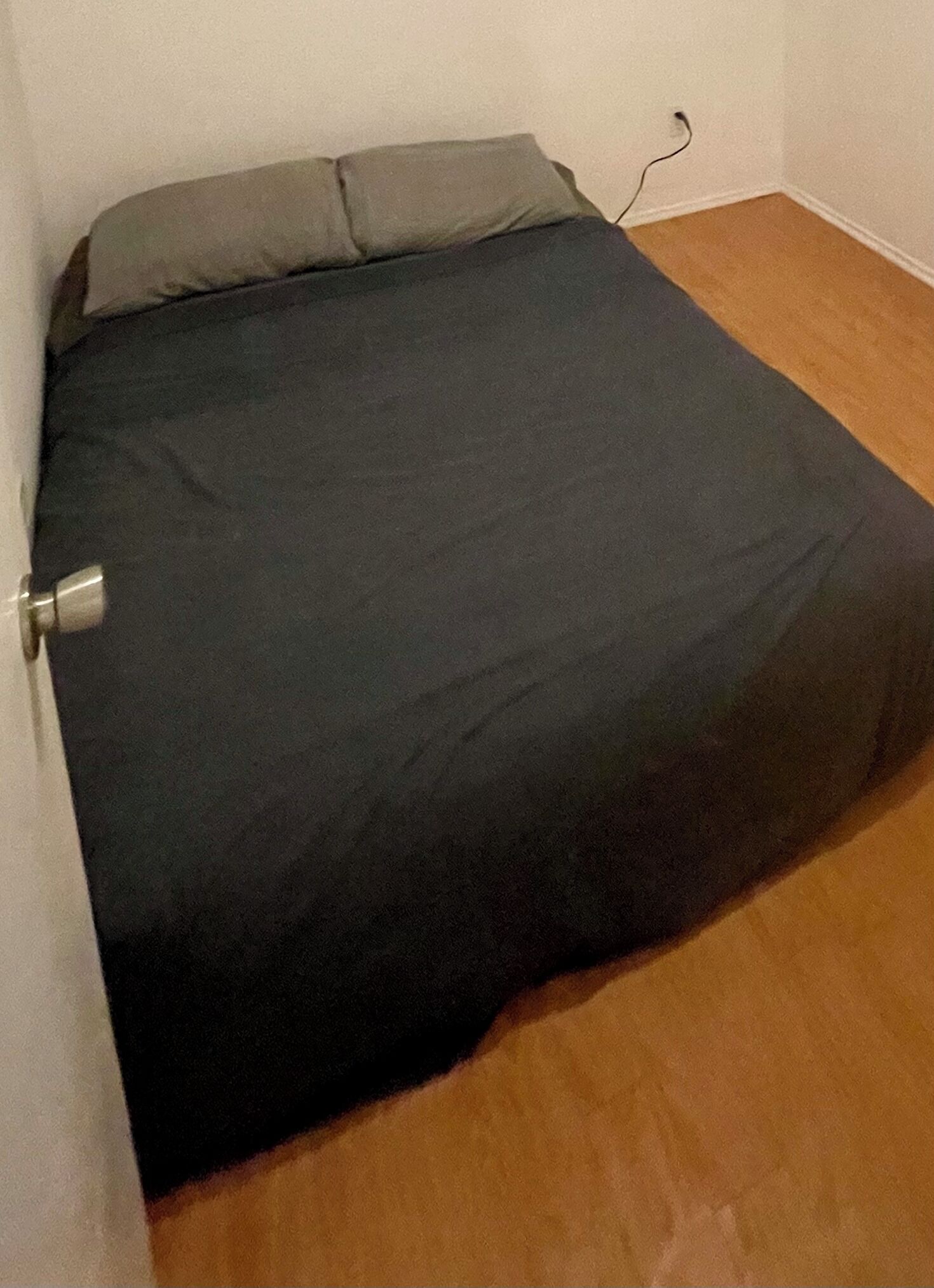 Une petite pièce faiblement éclairée, avec un lit simple, une prise de courant visible et un plancher en bois.