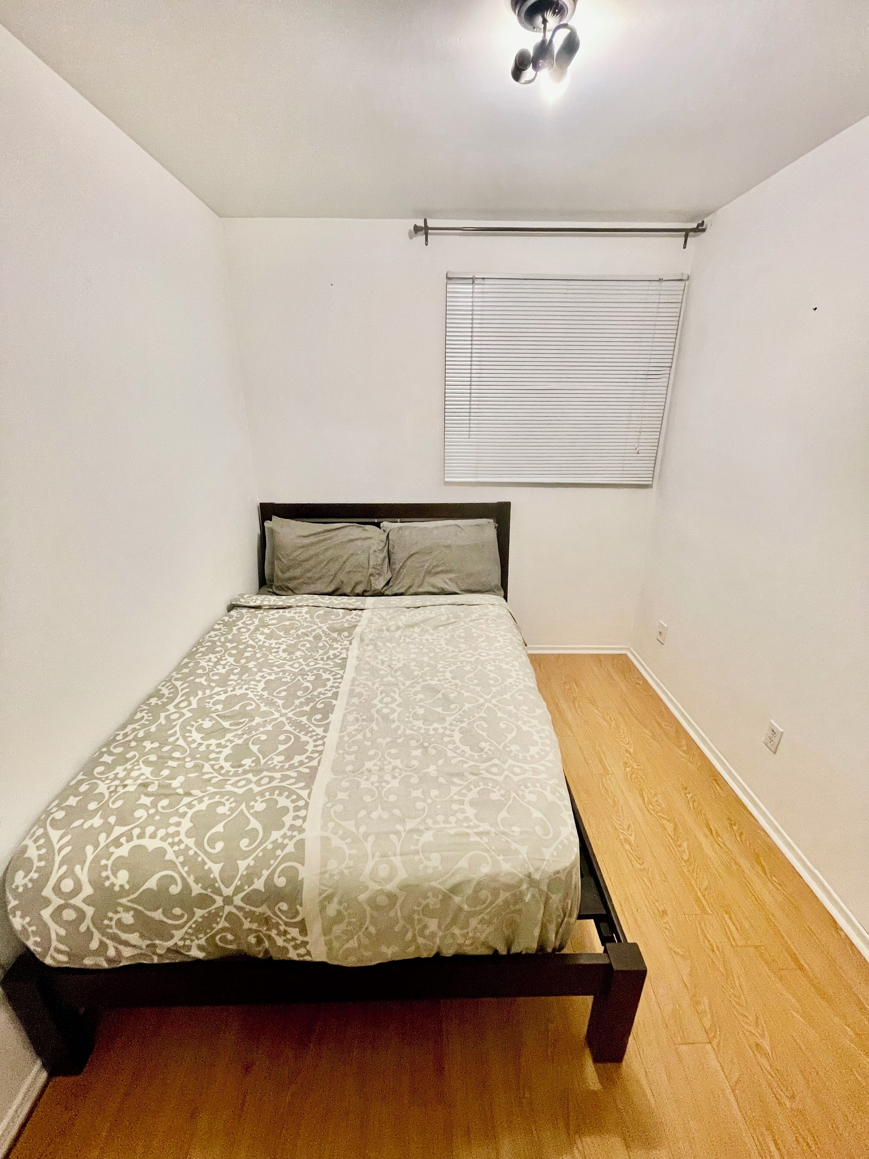 Une chambre à coucher avec un lit, un plancher en bois, des murs blancs, une fenêtre avec des stores et un luminaire au plafond.